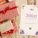 Karta podarunkowa o wartości 300 zł leżąca na stoliku obok zapakowanych prezentów. Karta ma jasne tło z różowym ozdobnym marginesem, widoczny napis BON PODARUNKOWY oraz logo DARYmex. W tle kolorowe, wzorzyste opakowania z czerwonymi i białymi motywami.