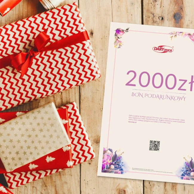 Bon podarunkowy o wartości 2000 zł na jasnym tle, z widocznym logo Darymex. Złożony na drewnianym stole, obok prezentów w czerwono-białą zygzakowatą i złotą ozdobą. Napisy w kolorze fioletowym. W tle różnokolorowe papierowe opakowania.
