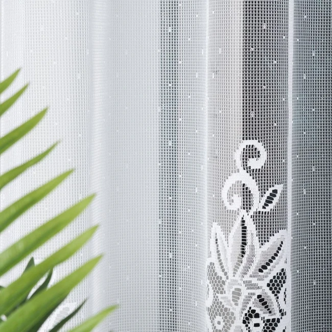 Firana gotowa Berenika w kolorze białym, wykonana z żakardowego materiału, o wymiarach 300x160 cm. Z detalami w postaci wzoru roślinnego. Kompozycja z boku, elementy tła z zielonymi liśćmi roślin. Firana rozłożona, wisząca, z delikatną teksturą.