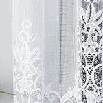 Firana żakardowa Berenika w kolorze białym o wymiarach 300x160 cm, zawieszona w pionie, z delikatnym, koronkowym wzorem. Tło proste, na jasnym, neutralnym tle, podkreślającym detale faktury i wzorów.