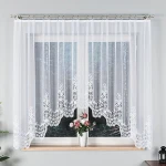 Firana gotowa Berenika żakardowa biała, o wymiarach 300x160 cm, z subtelnym wzorem w dolnej części. Zawieszona na karniszu, widoczne przezroczystości. Otoczenie: jasna ściana i kwiatek w doniczce na parapecie.