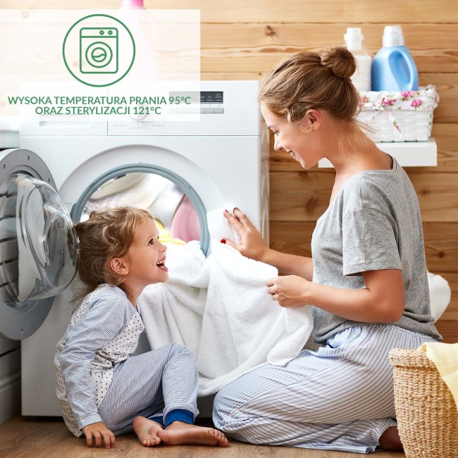 Kobieta w szarym T-shircie i spódnicach w paski w pozycji klęczącej, podaje dziecku w piżamie z kropkami białą ręcznik z pralki. Tło drewniane z detergentami w koszyku. Ujęcie frontalne, ukazujące radość dziecka i interakcję z dorosłym.