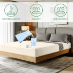Ochronne prześcieradło z gumką 160x200 w kolorze kremowym, rozłożone na łóżku. Na materiale widoczna woda, co ilustruje odporność na wilgoć. W tle nowoczesna sypialnia z szaro-brązowymi elementami. Kąt ujęcia frontalny z widokiem na poduszkę i stelaż.