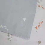 Pościel satyna bawełniana 220x200 Luxury Premium Perimelis w odcieniach bieli i szarości z delikatnymi kwiatowymi wzorami. Zbliżenie na poszewkę, leżącą na podłożu, z widocznymi motywami roślinnymi w ciepłych kolorach.