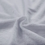 Poszewka frotte 40x40 w kolorze popiel jest rozłożona, ukazując teksturę miękkiego i przyjemnego materiału. Kąt ujęcia to zbliżenie, eksponujące detale splotu tkaniny. Tło jest jasne i uniwersalne, co podkreśla cechy poszewki.