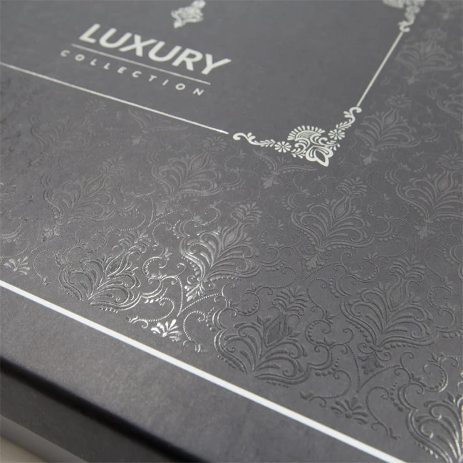 Opakowanie pościeli wykonane z czarnego kartonu z wypukłym wzorem roślinnym i napisem LUXURY COLLECTION. Widok na górną część pudełka, które jest otwarte. Biały pasek z logo umieszczony na krawędzi.