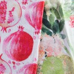 Ścierka kuchenna z motywem owoców tropikalnych, kolorowa z wzorem granatów oraz kolorowych kwiatów, wykonana z miękkiego materiału. Widok zbliżeniowy na dwa kawałki ścierki, z lekką teksturą tkaniny i wykończeniem wzdłuż krawędzi.