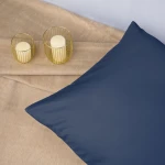 Poszewka satynowa 50x80 w kolorze chabrowym leży na jasnobeżowym tle. Ujęcie z boku pokazuje gładką fakturę materiału. Obok znajdują się dwa świeczniki w złotych osłonach z białymi świecami, tworząc elegancką aranżację.