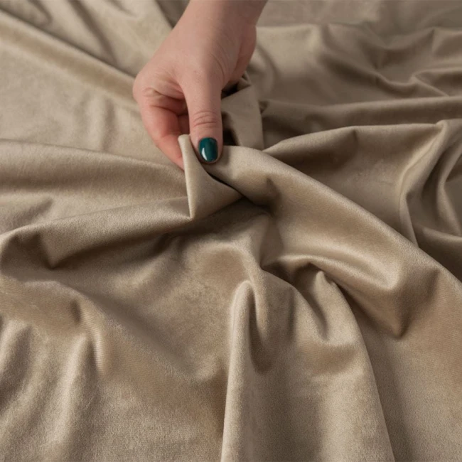 Zasłona gotowa welwetowa w kolorze ciemno beżowym, z miękkiej tkaniny Velvi. Zbliżenie na materiał, uchwycony w ręce, ukazuje gładką fakturę i naturalny blask tkaniny. Zasłona jest lekko pofalowana, prezentując swoje właściwości materiałowe.