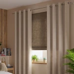Zasłona gotowa welwetowa 140x260 cm w ciemno beżowym kolorze, wisząca na przelotkach. Gładka, miękka tkanina z teksturą. Ujęcie frontalne, w otoczeniu minimalistycznej aranżacji wnętrza z roślinami i książkami w tle.