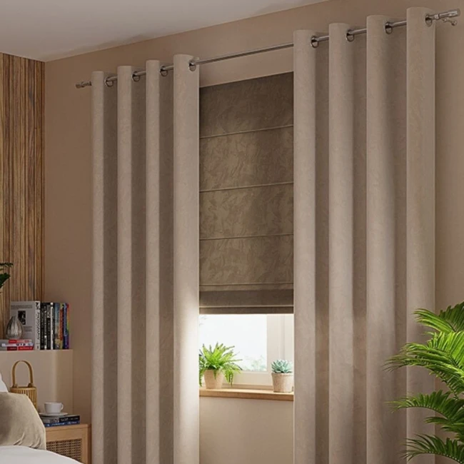 Zasłona gotowa welwetowa 140x260 cm w ciemno beżowym kolorze, wisząca na przelotkach. Gładka, miękka tkanina z teksturą. Ujęcie frontalne, w otoczeniu minimalistycznej aranżacji wnętrza z roślinami i książkami w tle.