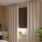 Zasłona welwetowa w kolorze kremowym, o wymiarach 140x260 cm, zawieszona na przelotkach. Tkanina ma miękką teksturę, a zasłona jest rozłożona w pionie. W tle widoczna jest roleta w odcieniu brązowym oraz rośliny doniczkowe.