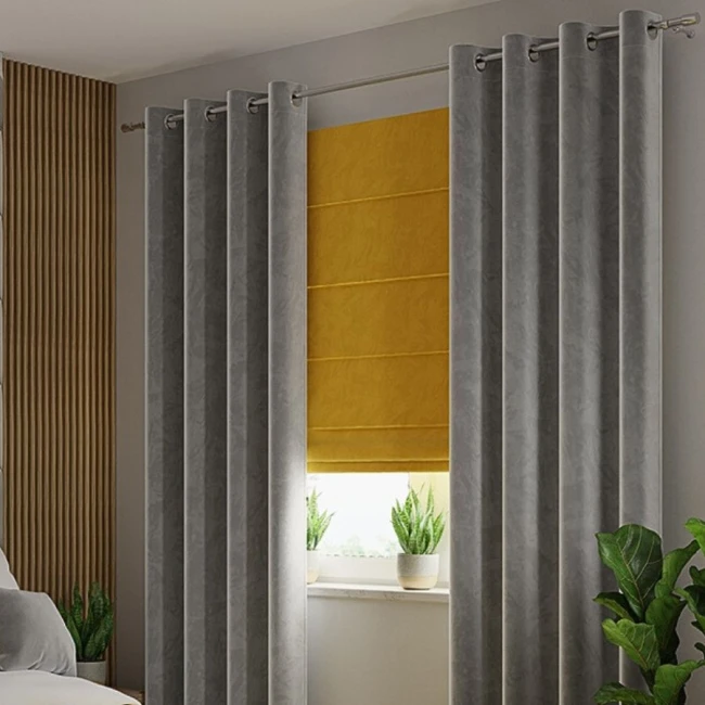 Zasłona welwetowa w kolorze szarym, rozmiar 140x260 cm, zawieszona na przelotkach. Produkt z miękkiej tkaniny, ukazany frontalnie w aranżacji wewnętrznej obok żółtej roletki. Tło neutralne, jasne z roślinami doniczkowymi na parapecie.
