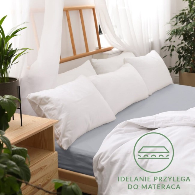 Prześcieradło jersey z gumką 160x200 w kolorze popielatym, leżące na materacu z poduszkami w białych poszewkach. Widoczny detal o logo IDEALNIE PRZYLEGA DO MATERACA na dolnym rogu. Tło w jasnym, przytulnym wnętrzu z roślinnością.