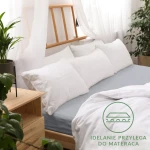 Prześcieradło jersey z gumką 180x200 w kolorze popielatym, gładkie, dobrze przylegające do materaca. Widoczne na łóżku z białymi poduszkami i narzutą. Otoczenie domowe, drewniane elementy, rośliny w tle.