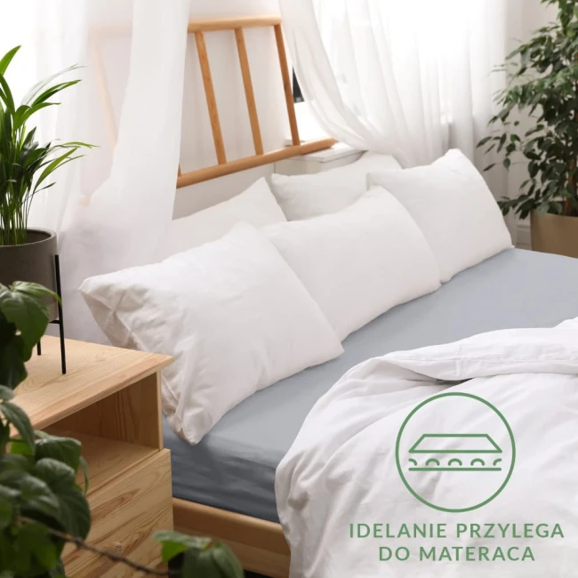 Prześcieradło jersey z gumką 200x220 w kolorze popielatym rozłożone na łóżku, otoczone białymi poduszkami. Ujęcie z boku ukazuje gładką fakturę materiału.