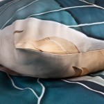 Pościel satyna bawełniana 220x200 Satynlove Premium w odcieniach złotej i turkusowej z delikatnym wzorem liści. Poduszka leży na miękkim tle z fakturą. Widać gładkie wykończenia i elegancki nadruk, co nadaje jej nowoczesny wygląd.