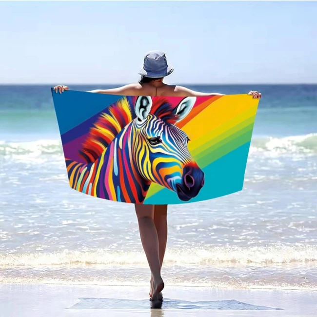 Ręcznik plażowy 100x180 z mikrofibry, kolorowa zebra na tle tęczowych pasów. Ułożony w poziomie, trzymany przez osobę w kapeluszu, stojącą nad brzegiem morza. Szybkoschnący materiał, duży rozmiar idealny na plażę.