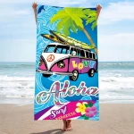Ręcznik plażowy 100x180 cm, z mikrofibry, w kolorze niebieskim z grafiką surfingu, kwiatami i napisem Aloha Love. Złożony w pionie, trzymany przez osobę na plaży, z falami i palmami w tle. Szybkoschnący materiał idealny na plażę.