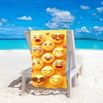 Ręcznik plażowy 100x180 cm z mikrofibry w intensywnym żółtym kolorze, z grafiką różnych emotikonek. Leży na leżaku na plaży, w tle widoczna błękitna woda i chmury. Szybkoschnący materiał, duży rozmiar do użytku na plaży.
