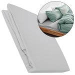 Prześcieradło flanelowe z gumką 140x200 w kolorze jasno szarym, wykonane z miękkiego materiału. Widoczne złożenie prześcieradła, które leży na białym tle. W tle fragment łóżka z pościelą w kolorze szaro-zielonym, poduszkami oraz narzutą.