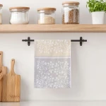 Ścierka bawełniana kuchenna 50x70 w kolorze kremowym z białym wzorem gałązek, zawieszona na czarnym wieszaku. Ułożona w pionie, w tle widoczne szklane słoiki na półce, co tworzy kuchenny kontekst aranżacyjny.