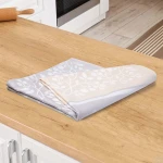 Ścierka bawełniana kuchenna 50x70 w kremowej kolorystyce z delikatnym wzorem gałązek. Ułożona na drewnianym blacie kuchennym, w stanie leżącym. Tło w kuchennej aranżacji z widocznym piekarnikiem w tle.