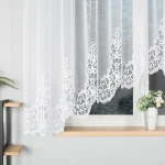 Firana gotowa BERENIKA żakardowa, o wymiarach 400x160 cm, w kolorze białym. Zawieszona na oknie, z haftowanym wzorem na dolnej krawędzi. Tło neutralne, z wazonem oraz rośliną w pobliżu. Przez firanę prześwituje światło, nadając przytulności wnętrzu.