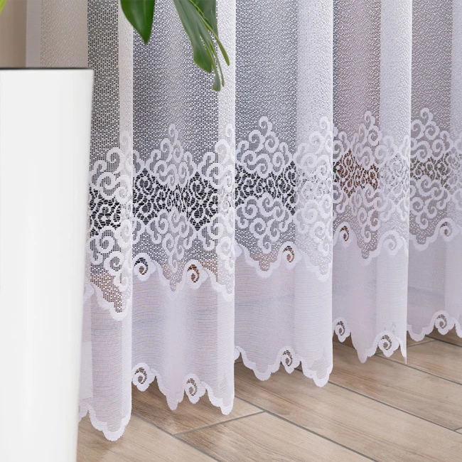 Firana gotowa MODESTA żakardowa, biała, o wymiarach 200x250 cm. Ujęcie z boku, wisząca, z delikatnym wzorem w dolnej części, z wykończeniem w falbanki. Transparentna tkanina dobrze przepuszczająca światło, na tle jasnej podłogi.