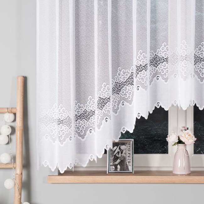 Firana gotowa MODESTA, żakardowa, w kolorze białym, o wymiarach 300x160 cm, zawieszona na oknie. Widoczne eleganckie, faliste wzory u dołu. Tło w stonowanej szarości, z okna i dekoracjami na parapecie, w tym zdjęciem i wazonem z kwiatami.