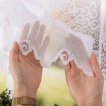Firana gotowa MODESTA, żakardowa, biała, o wymiarach 500x160 cm, z dekoracyjnym, falistym wykończeniem na dole. Prezentowana w dłoniach, część materiału z widoczną fakturą w tle. Ujęcie z bliska, akcentujące szczegóły wzoru.