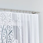 Firana żakardowa w kolorze białym o wymiarach 300x160 cm, prezentująca delikatny wzór roślinny. Ujęcie frontalne z góry, zwisająca na karniszu. Tło w jasnym odcieniu, co podkreśla jej jasność i szczegóły.