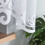 Firana gotowa POLA żakardowa, 300x160 cm, biała, zawieszona na karniszu. Zbliżenie na dolną krawędź zasłony z haftem w kształcie liści. Tło w odcieniu szarości, obok widoczna zielona roślina w doniczce. Tekstura w finezyjny wzór, lekko przezroczysta.