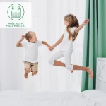 Ochraniacz na materac 90x160 Robson w kolorze białym, umieszczony na łóżku. Na zdjęciu dzieci w białych ubraniach skaczą radośnie, trzymając się za ręce. Tło z jasnymi zasłonami i zielonymi firankami.
