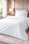 Pościel hotelowa bawełniana 220x200 w białe pasy 1 cm, złożona na łóżku. Poduszka z identycznym wzorem, widoczne delikatne fakturki. Tło w stonowanej kolorystyce wnętrza z rośliną i drewnianym stolikiem.