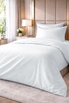 Pościel hotelowa bawełniana 140x200 w kolorze białym, z delikatnym wzorem w pasy o szerokości 1 cm. Złożona, leżąca na łóżku, z poduszką w komplecie. Tło w jasnym, stonowanym kolorze z elementami roślinnymi w aranżacji.
