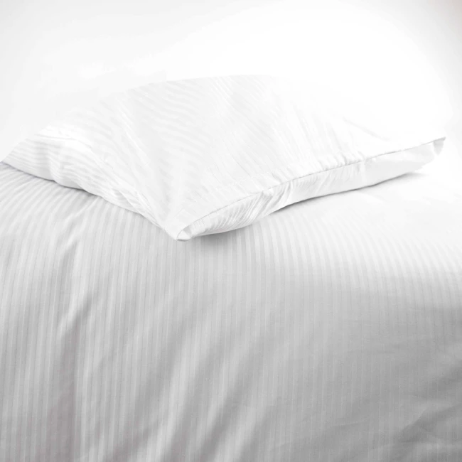 Pościel hotelowa bawełniana 220x200 w pasy 4-mm, biała, leżąca na podłożu. Widoczna poszewka na poduszkę z delikatnym przeszyciem. Faktura tkaniny gładka, z subtelnym połyskiem, na jednolitym tle. Rozmiar poduszki idealnie dopasowany do pościeli.