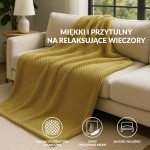 Koc żakardowy 150x200 w kolorze oliwkowym, z fakturą w formie drobnych, regularnych wzorów, leży na szarej, kanapie. W tle widoczna lampa na stoliku, co tworzy przytulny klimat aranżacji wnętrza.
