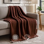 Koc żakardowy 150x200 w kolorze mlecznej czekolady, z fakturą w formie drobnych, regularnych wzorów, leży na szarej, kanapie. W tle widoczna lampa na stoliku, co tworzy przytulny klimat aranżacji wnętrza.