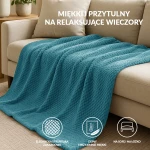 Koc żakardowy 150x200 w kolorze jasnym turkusowym, z fakturą w formie drobnych, regularnych wzorów, leży na szarej, kanapie. W tle widoczna lampa na stoliku, co tworzy przytulny klimat aranżacji wnętrza.