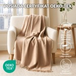 Koc bawełniany 150x200 cm w jasnym beżu, z frędzlami na dolnej krawędzi, leży na jasnej kanapie. W tle widać okno oraz dekoracyjne zasłony. Koc ma certyfikat Oeko-Tex, co potwierdza jego jakość i bezpieczeństwo.