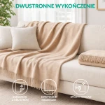 Koc bawełniany w jasnym beżu, o wymiarach 150x200 cm, z frędzlami. Układ leżący na kanapie, z dwustronnym wykończeniem i estetycznymi krawędziami. W tle poduszki w jasnym kolorze i neutralne tło wnętrza.