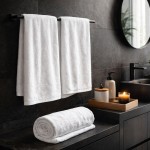 Biały ręcznik hotelowy 50x100 cm, gładki, o gramaturze 400 g/m², leży zwinięty na blacie, obok dwóch ręczników wiszących na wieszakach. W tle widoczne akcesoria łazienkowe oraz lustro. Tło ciemne, minimalistyczne.