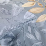 Pościel bawełniana 220x200 w kolorze niebieskim z beżowym wzorem liści. Zbliżenie na poszewkę z delikatnym nadrukiem, stworzona z gładkiego materiału. Widoczne liście w różnych odcieniach, leżąca na płaskiej powierzchni.