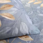 Pościel bawełniana 220x200 Sandleaf w niebieskim kolorze z beżowymi liśćmi. Wzór liściowy na poszewkach i narzucie, z miękką, gładką fakturą materiału. Pozycja leżąca, z bliskim ujęciem detali.