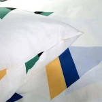 Pościel bawełniana 160x200 w kolorze białym z kolorową, geometryczną grafiką. Zbliżenie na poszewkę z widocznymi zielonymi, niebieskimi i żółtymi wzorami na tle. Materiał gładki, o matowej fakturze, leżący na płaskiej powierzchni.