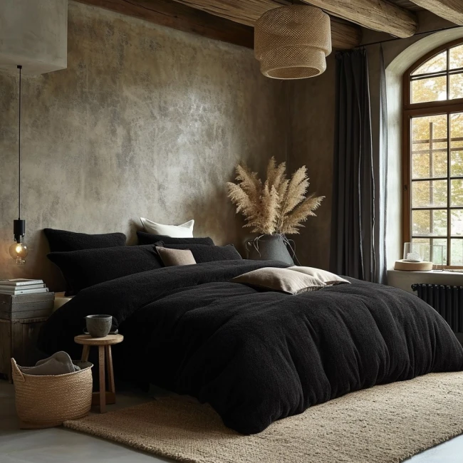 Poszewka frotte 40x80 w kolorze czarnym, wykonana z miękkiego materiału, leżąca na łóżku z poduszkami. W tle widoczna dekoracja roślinna oraz oświetlenie sufitowe. Całość w stonowanej aranżacji sypialni.