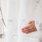 Firana gotowa KOKARDKA w kolorze białym, haftowana srebrnymi wstążkami, z ołowianką, na taśmie. Widoczna ręka dotykająca materiału. Wymiary: 140 x 250 cm. Tło jasne, proste, co podkreśla detale haftu.