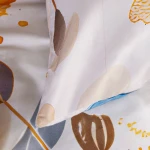 Pościel satynowa 200x200 cm w kolorze kremowym z niebieskimi motylami. Zbliżenie na poduszkę z delikatnym wzorem w odcieniach brązu i błękitu. Materiał gładki, błyszczący. Produkt leżący na płaskiej powierzchni. W tle neutralne, jasne kolory.