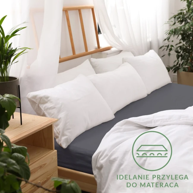 Prześcieradło jersey z gumką w kolorze antracytowym, leżące na łóżku, idealnie dopasowane do materaca, z widoczną białą pościelą i poduszkami. Tło w jasnych odcieniach, z drewnianym wezgłowiem i rośliną doniczkową w aranżacji sypialni.